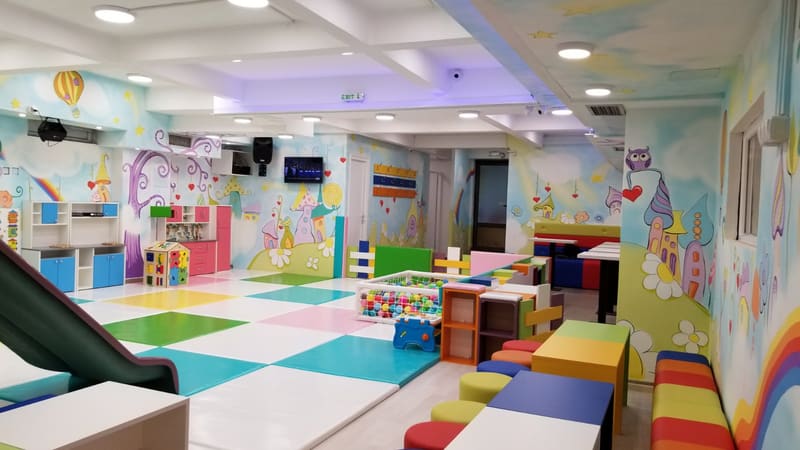 Igraonica sa mekim podom u boji, tapaciranim klupama, dečijim kuhinjama i oslikanim zidovima