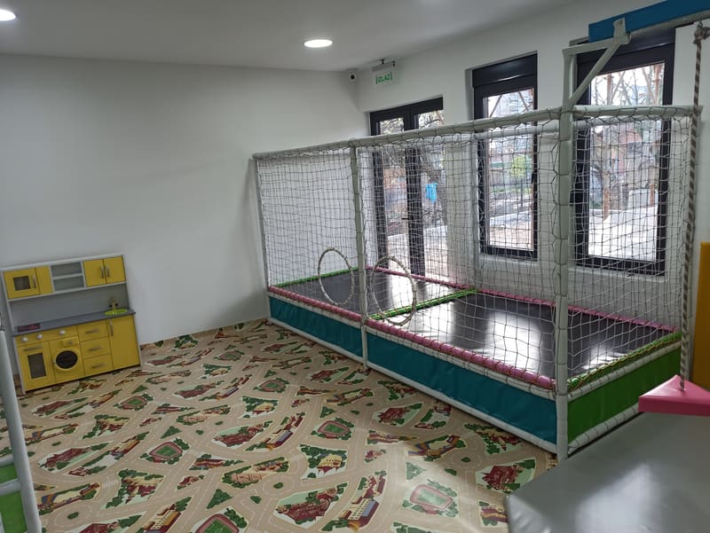 Trambolina u mrežastom kavezu sa obručima na ulazu, uz dečiju kuhinju i tepih sa ulicama